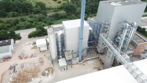 Luftaufnahme eines Biomassekraftwerkes (Steinbeis Energie Papenburg) mit Gebäuden, Schornstein, Rohrleitungen und Bauausrüstung, umgeben von Grünflächen und einer Straße. Finanzierung neue Rauchgasreinigung.