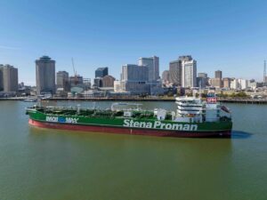 Green tanker ship ‘Stena Proman’ with ‘IMOIIMAX’ branding sailing through an urban harbor under clear skies, with a modern city skyline in the background.